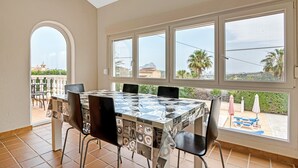 A dining area with a table and chairs, large windows with a view, and a ceiling light. - Villa 'Casa Leuki' with Sea/Mountain Views, Pool, Garden, Wi-Fi & AC (Calpe)
