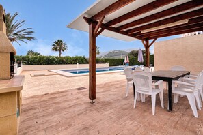 A covered outdoor area with a pool, white plastic chairs, a wooden table, and a pergola.