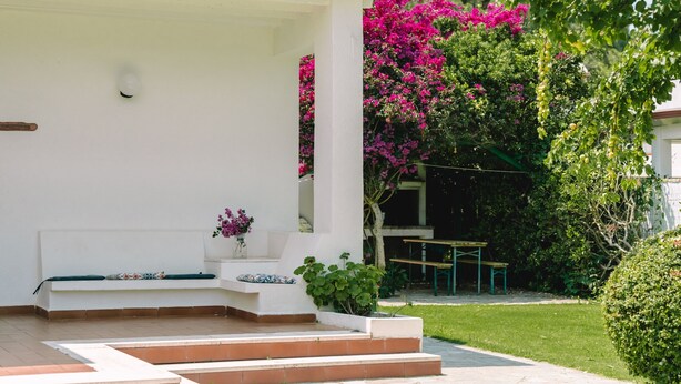 A white building with a covered porch, steps leading to a bench, and a garden with pink flowers and green shrubs. - Holiday Home "Villa Margherita" with 2 Private Terraces and Wi-Fi (Geremeas)