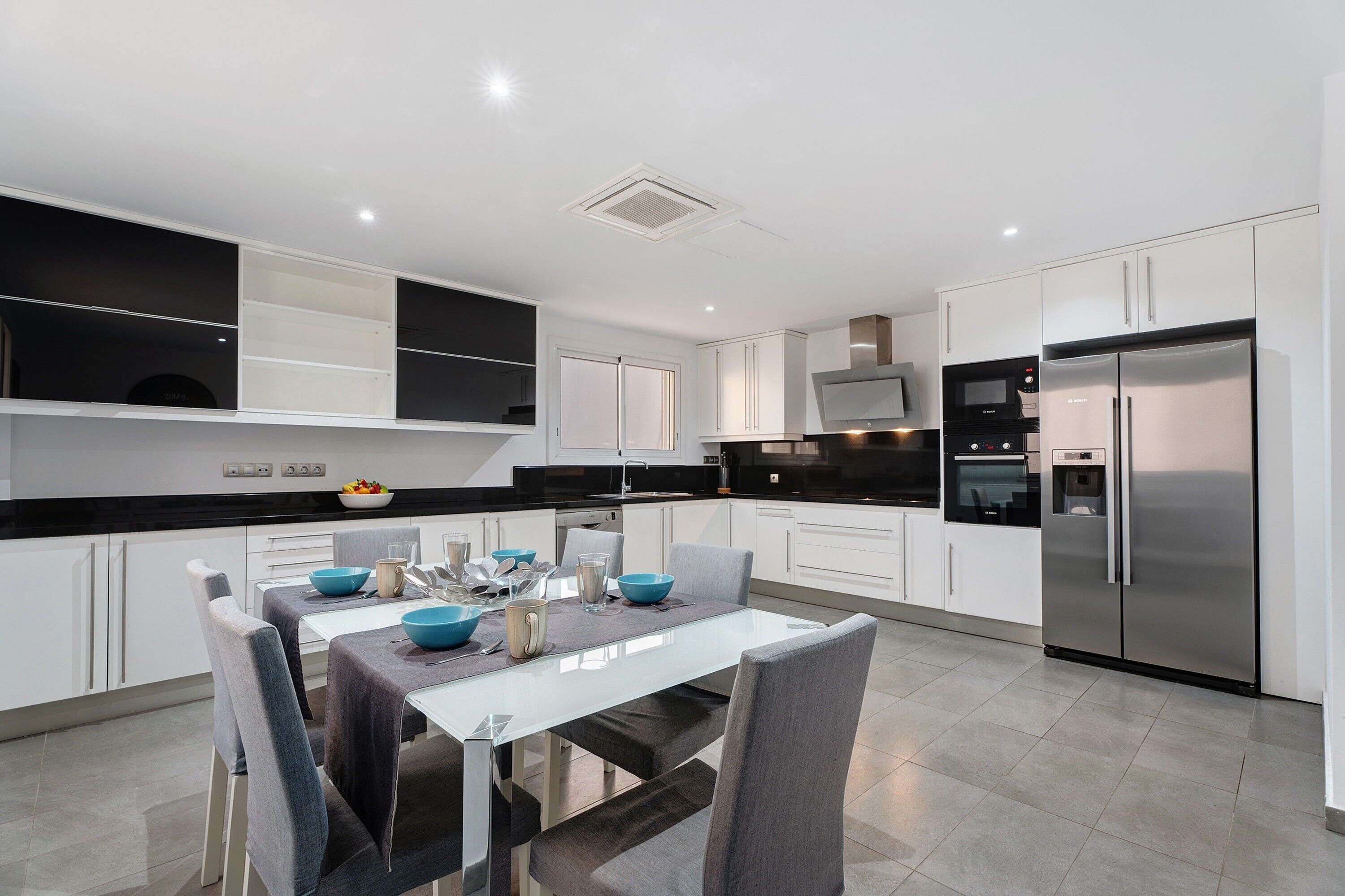 A modern kitchen with white cabinetry, a stainless steel refrigerator, and a dining area with a glass table and chairs.