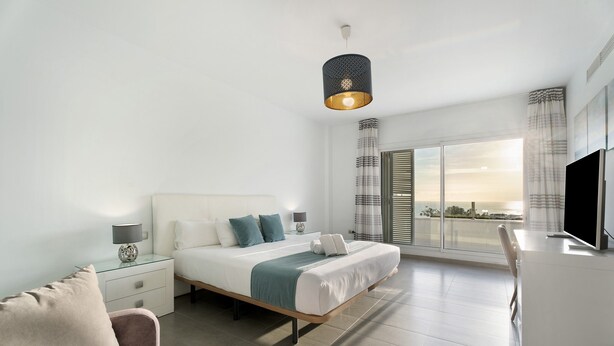 Une chambre d’hôtel moderne équipée d’un lit, de tables de chevet, d’un bureau, d’une chaise, d’une télévision et d’une grande fenêtre avec des rideaux.