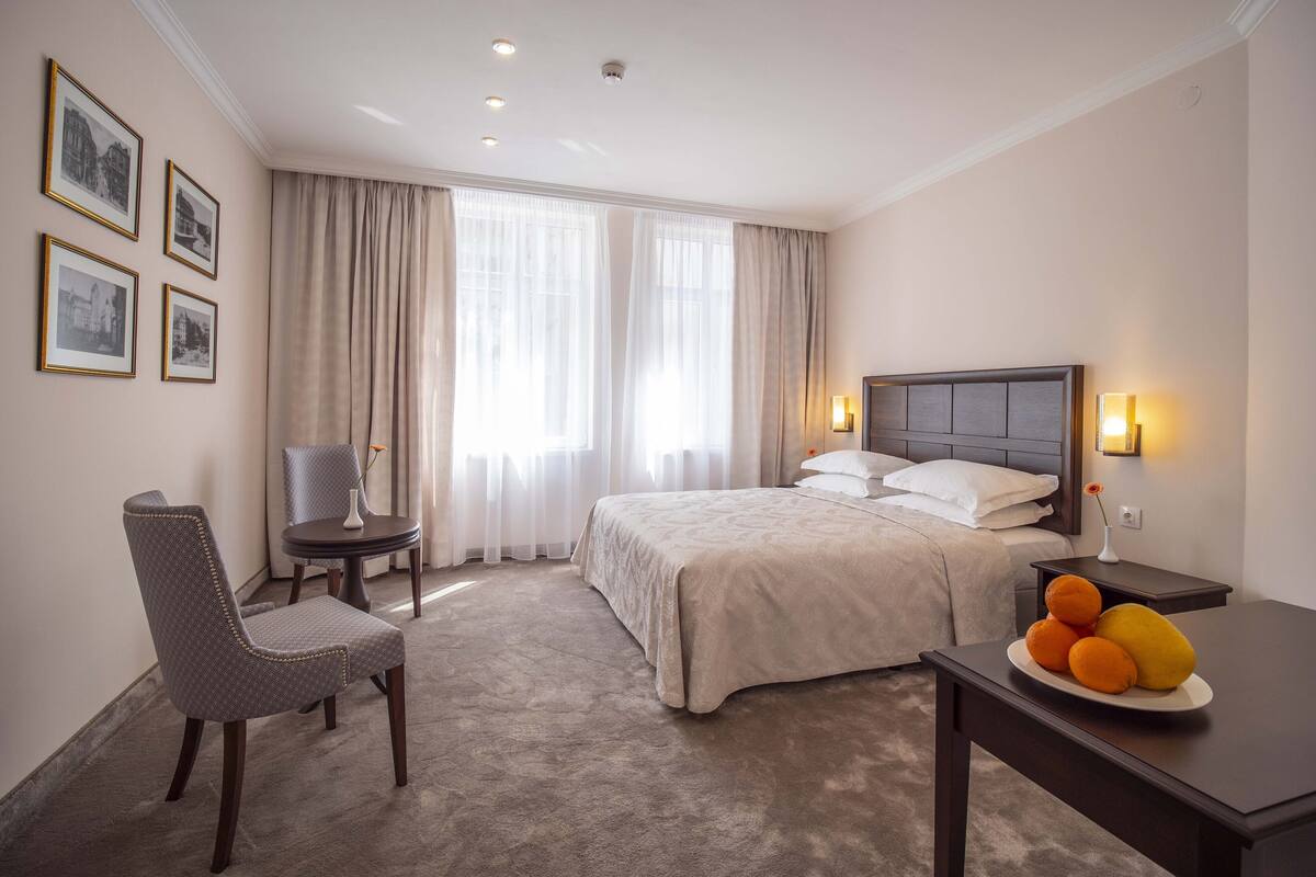 A hotel room with a bed, a chair, a table with fruit, and a window with curtains.