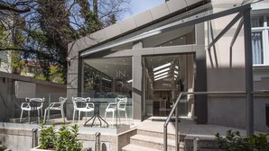 Un edificio moderno con acceso de vidrio, zona de estar al aire libre con sillas blancas y una escalera de piedra.