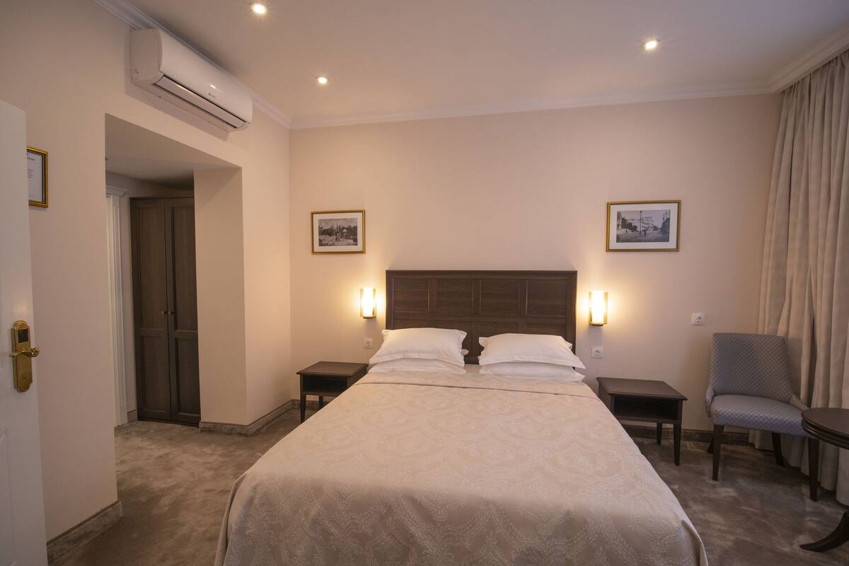 A hotel room with a large bed, two bedside tables, a chair, and an air conditioning unit on the wall.