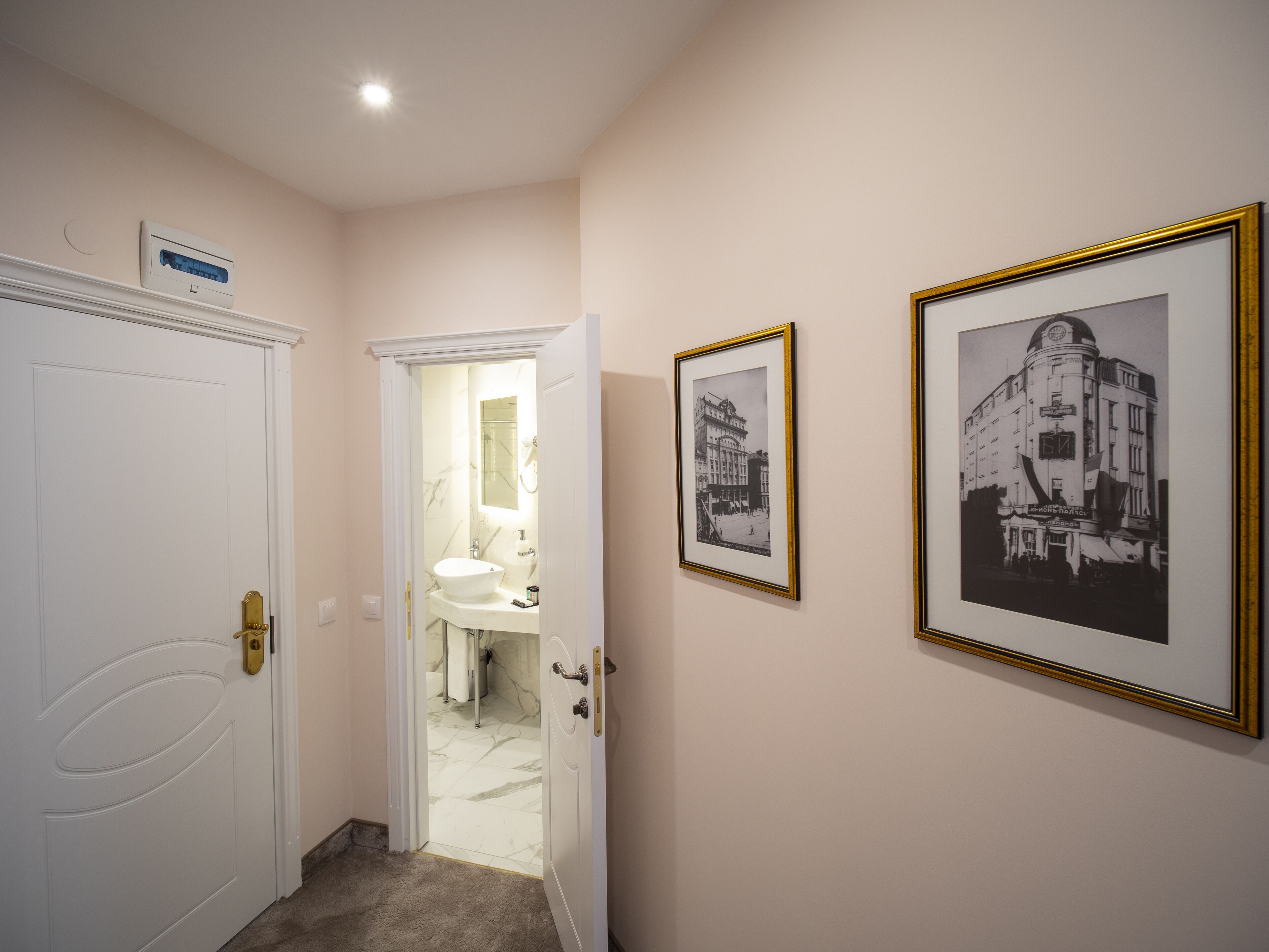 Een hal met een witte deur, een badkamer met een wastafel en spiegel, en twee zwart-wit foto's in lijsten aan de muur.