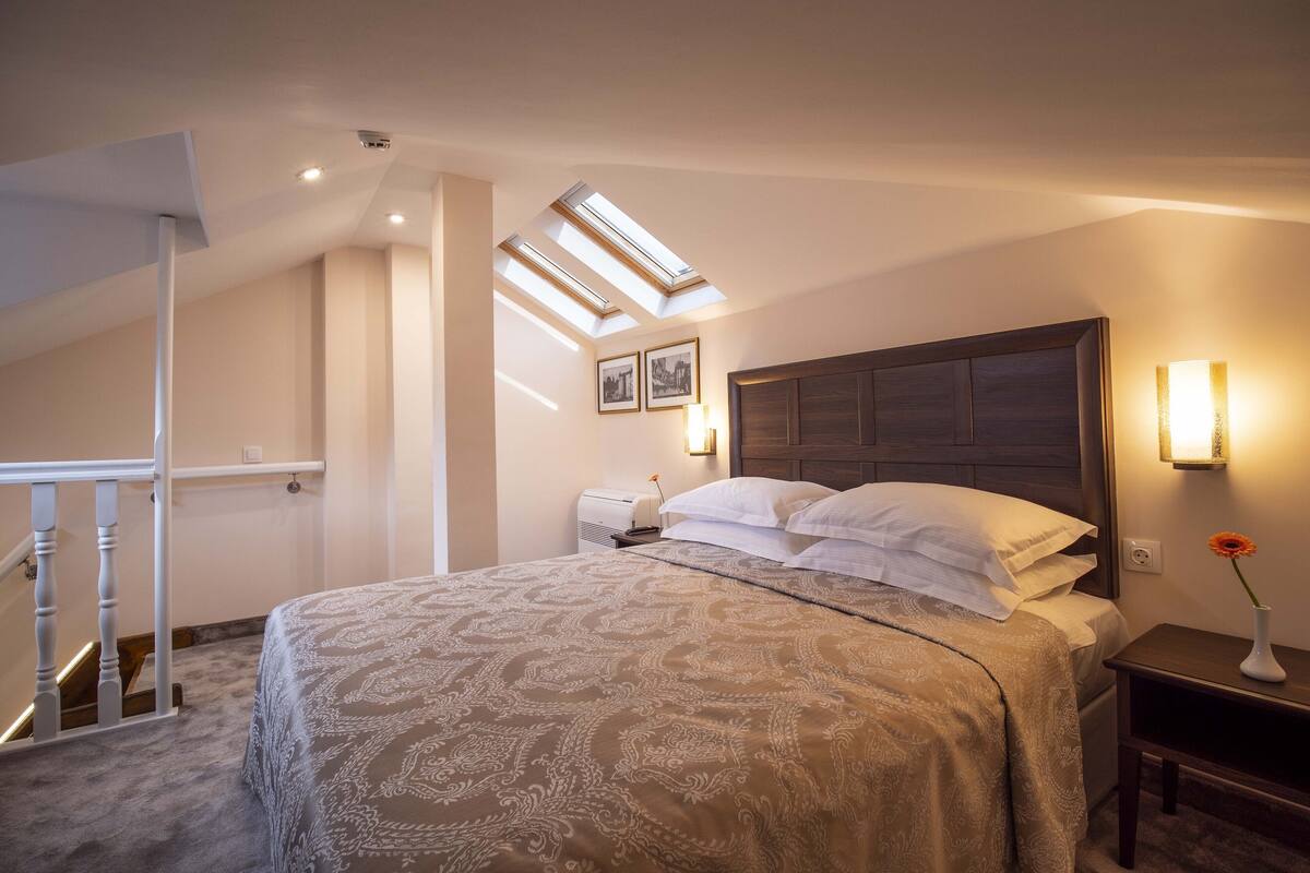 A bedroom with a large bed, a skylight, a nightstand, and a wall-mounted lamp.
