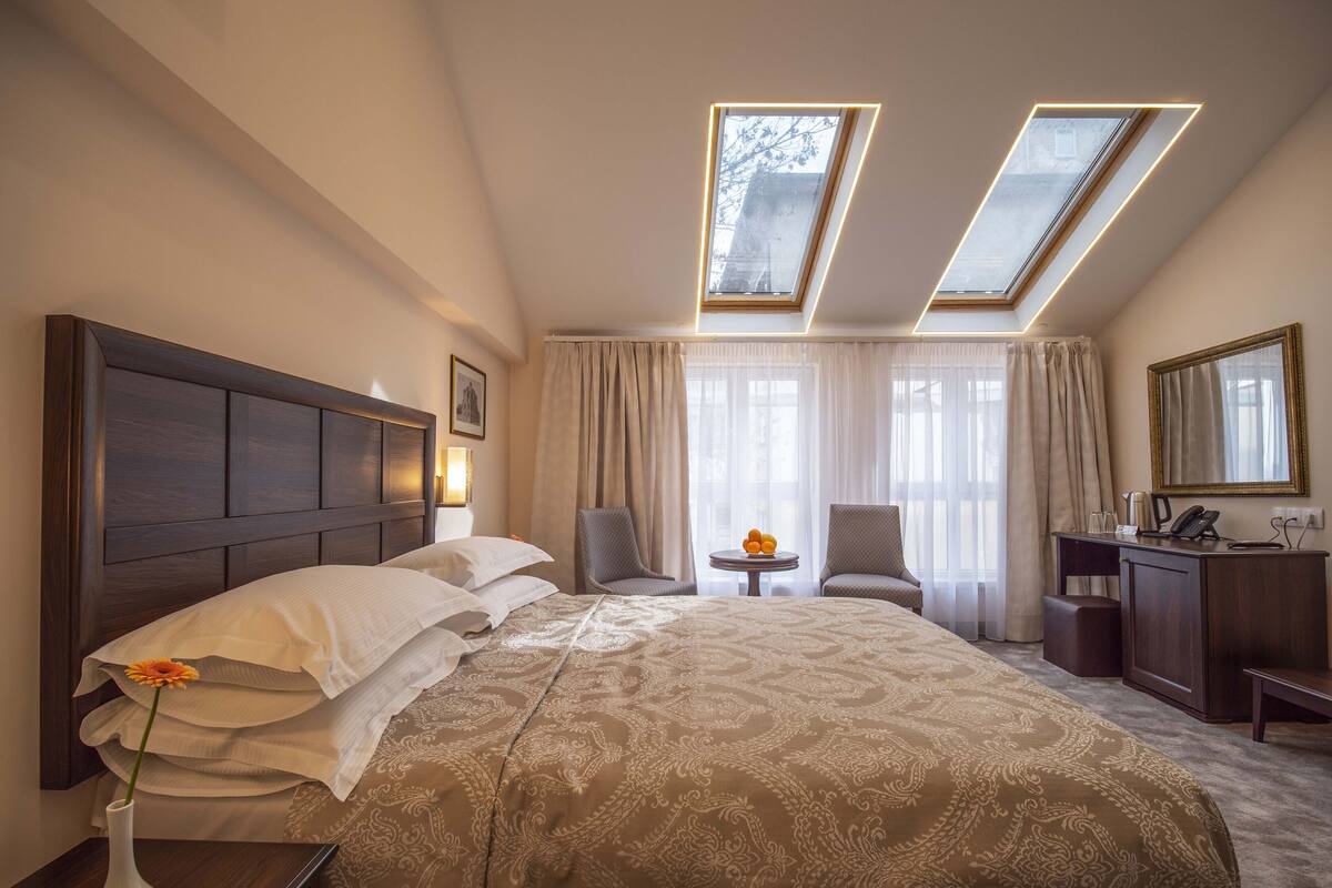 A hotel room with a large bed, two chairs, a desk, and a small table with a vase of flowers. There are skylights above the bed and a large window with curtains.