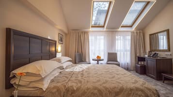 A hotel room with a large bed, two chairs, a desk, and a small table with a vase of flowers. There are skylights above the bed and a large window with curtains.