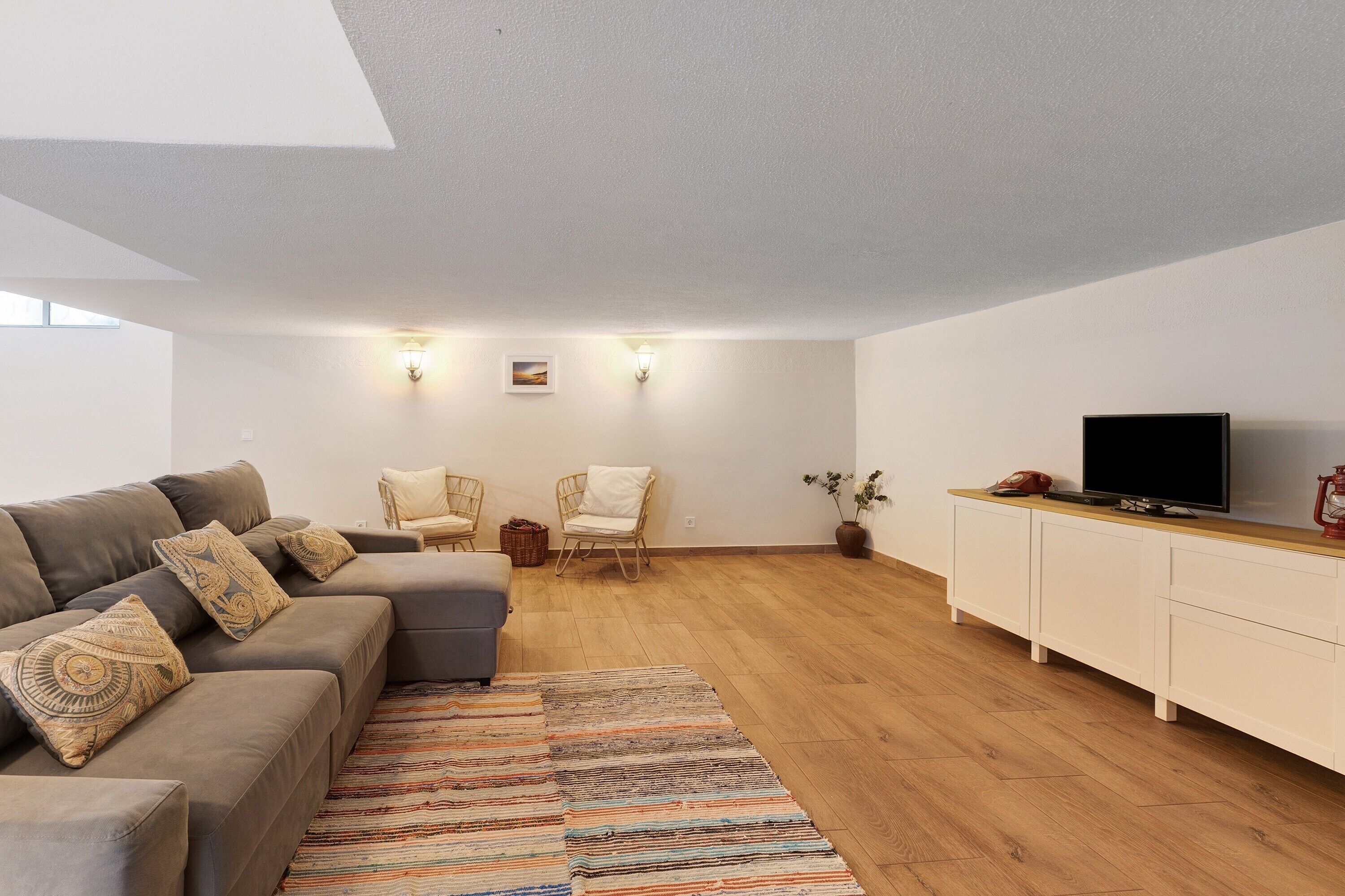 A modern living room with a grey sofa, patterned cushions, a wooden floor, a white cabinet, a television, and wall-mounted lights.