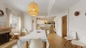 Ein Essbereich mit einem langen Tisch, weißen Stühlen, einem Kamin und einer Küche im Hintergrund.