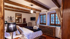 Ein Schlafzimmer mit Bett, Lampe, Fernseher, einem Fenster mit Vorhängen und einer Holzbalkendecke.