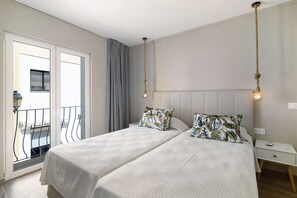 A double bed with patterned pillows, a bedside table with a lamp, and a view of a balcony with a railing.