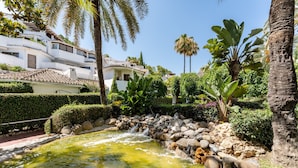 Un jardin paisible avec un étang, des palmiers et un bâtiment blanc doté d’un toit en tuiles.
