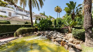 Un jardin paisible avec un étang, des palmiers et un bâtiment blanc doté d’un toit en tuiles.