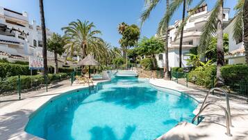 Une piscine à l’eau d’un bleu cristallin, entourée de palmiers et bordée d’un bâtiment blanc doté de balcons.