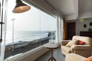 Een kamer met een groot raam met uitzicht op zee, een bank, een kleine tafel en een lamp.
