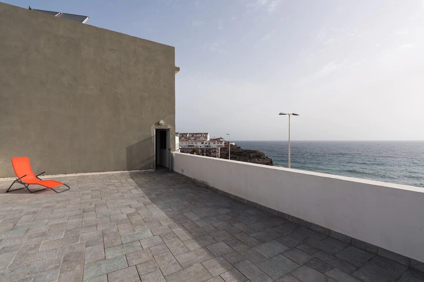 A balcony with a red chair, a view of the sea, and buildings in the background.