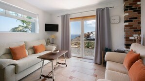 Un salon avec un canapé, un fauteuil et une table sur laquelle se trouve un saladier. Il y a un téléviseur accroché au mur, une horloge et une vue sur l’extérieur.