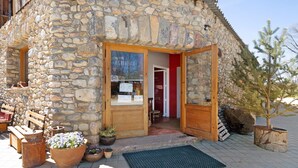 Un bâtiment en pierre avec une porte et des fenêtres en bois, un banc et des plantes en pot à l’extérieur.