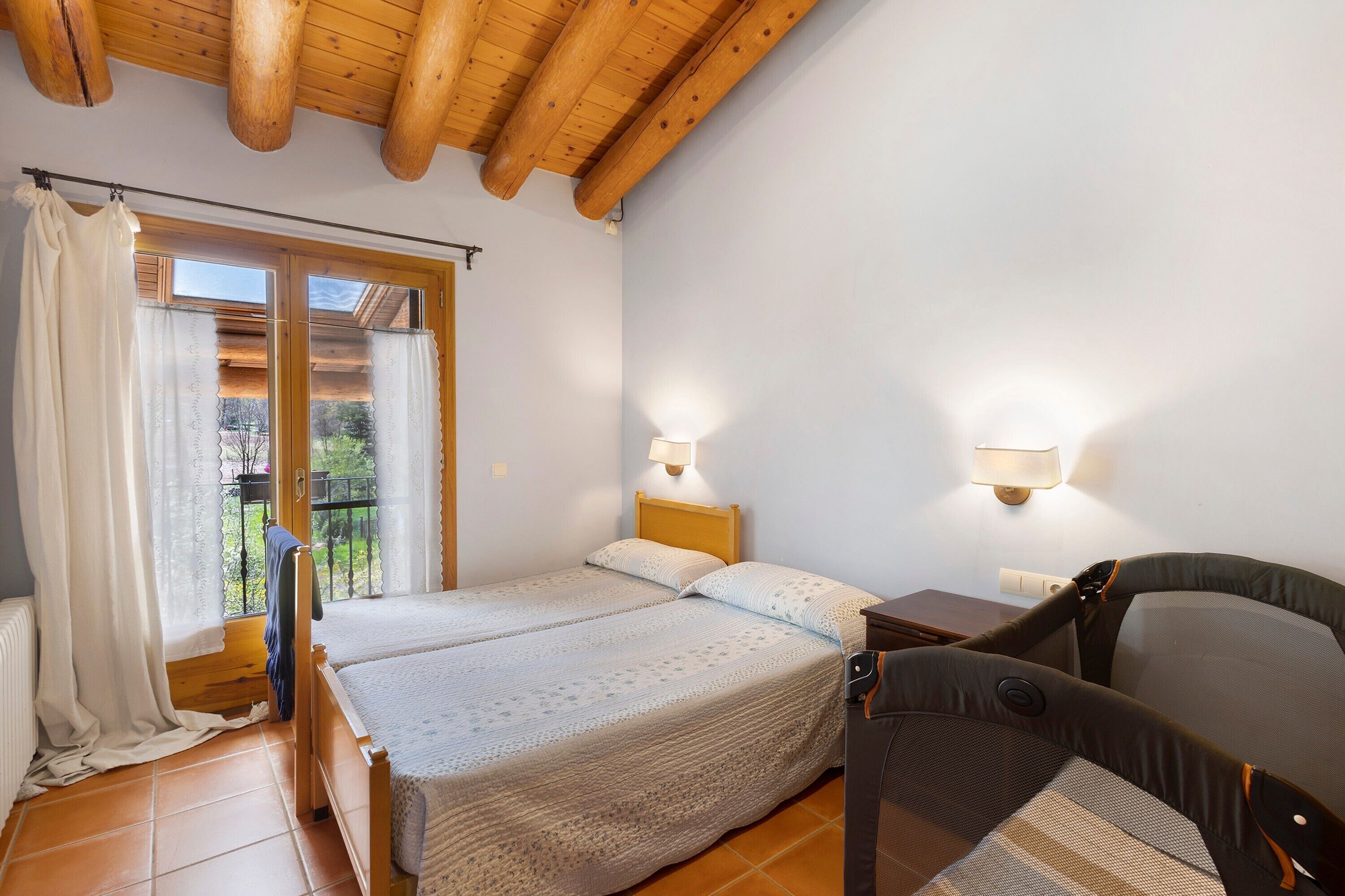 A room with two beds, a wooden ceiling, a balcony with a view, and two wall-mounted lights.