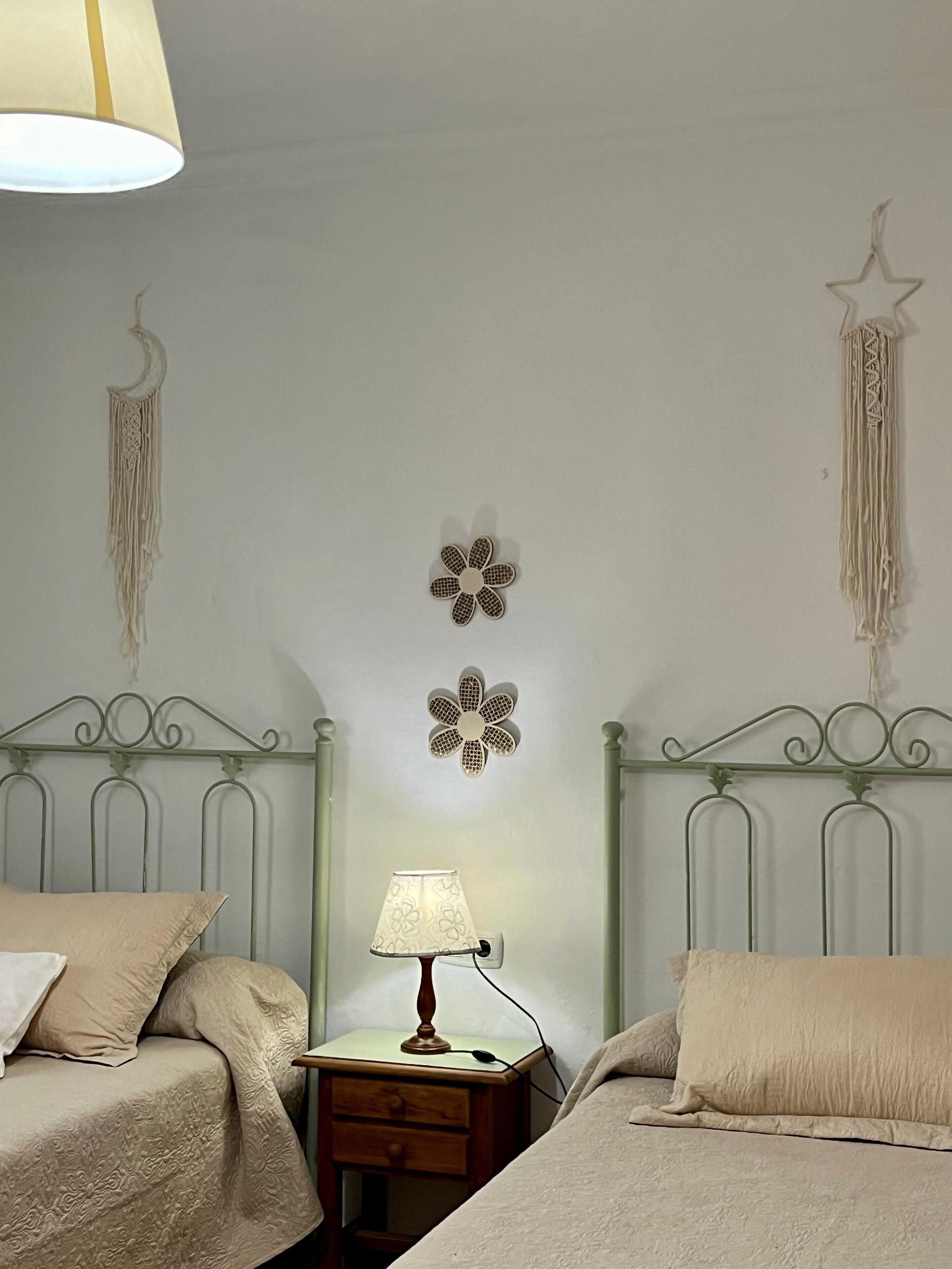 A bedroom with two beds, a nightstand with a lamp, and wall decorations including a flower and a hanging macrame.