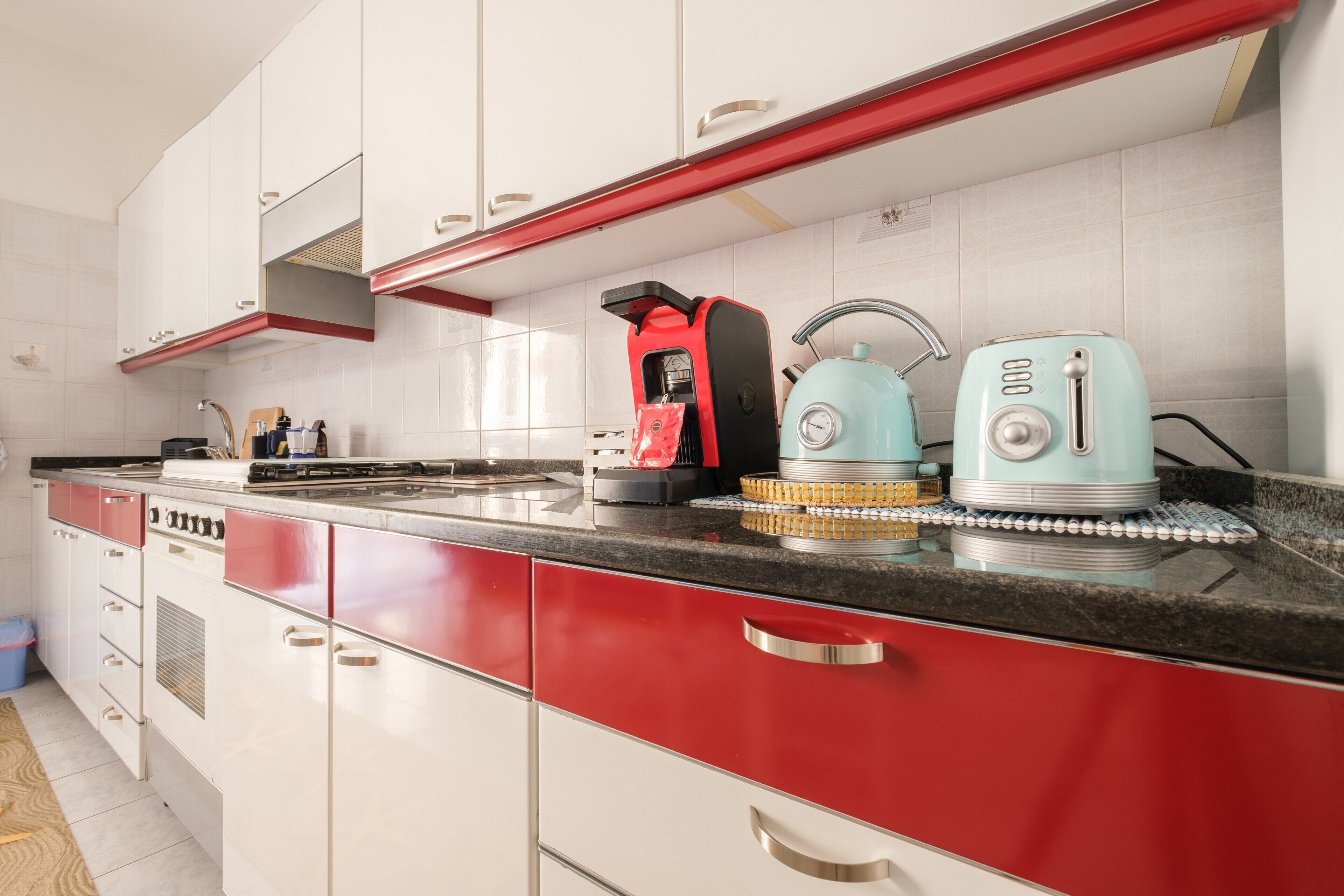Una cocina con armarios blancos, tiradores rojos, una cafetera negra y dos hervidores azules sobre la encimera.