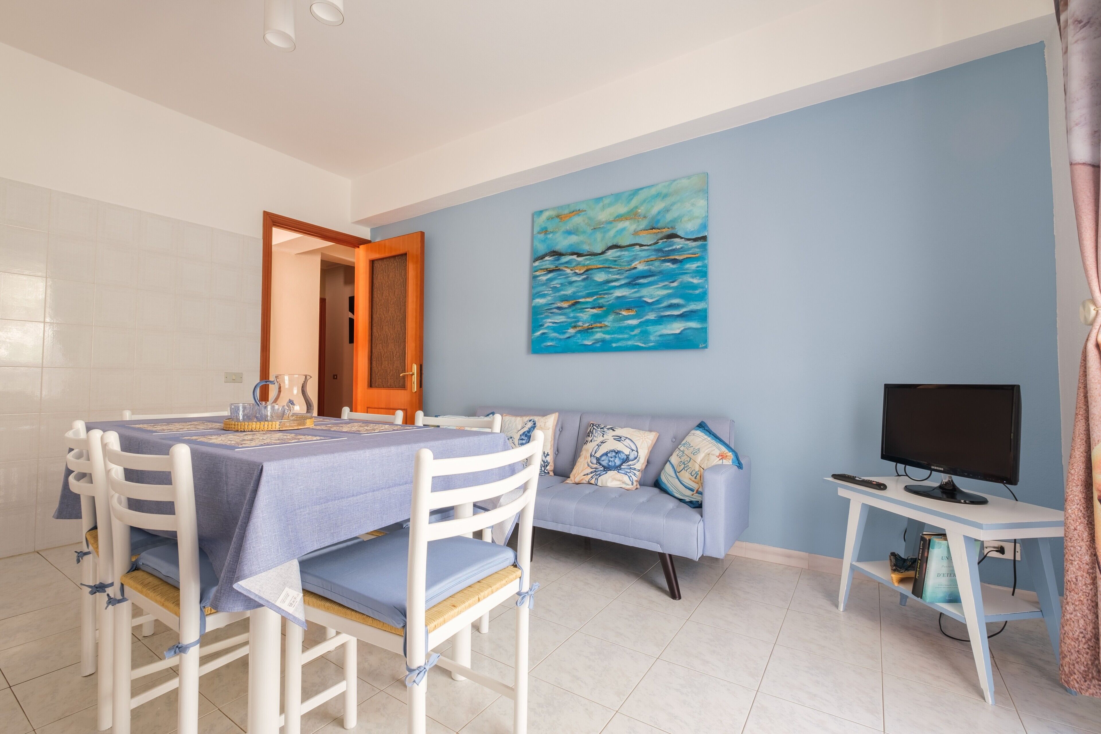 Un área de comedor con una mesa preparada para cuatro personas, un sofá, un televisor sobre un mueble bajo y un cuadro en la pared.