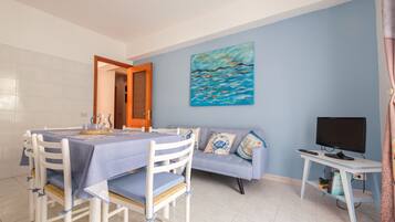 Un área de comedor con una mesa preparada para cuatro personas, un sofá, un televisor sobre un mueble bajo y un cuadro en la pared.