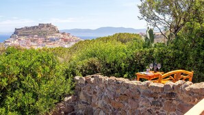 A stone wall with a wooden table and chairs, overlooking a coastal town with a fort on a hill.