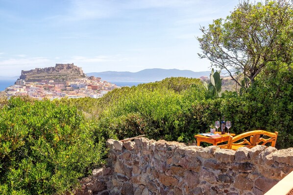 Mural de pedra com uma mesa e cadeiras de madeira, com vista para uma vila costeira e um forte numa colina.