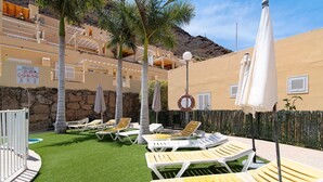 A pool area with lounge chairs and umbrellas, a stone wall, and a building with balconies.