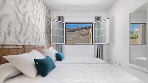 A bedroom with a large bed, two pillows, a window with curtains, and a view of a rocky landscape.