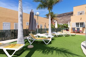 Une cour avec une pelouse artificielle, des chaises longues, des parasols et un palmier.