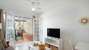 A living room with a TV on a white stand, a white sofa, a dining table with chairs, and a sliding glass door leading to a balcony.