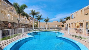 A swimming pool with a curved design, surrounded by a white fence, and adjacent to a building with balconies and palm trees.