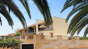 A modern two-story house with a balcony, a stone wall, and a palm tree in the foreground. - Vacation Home "Puerto Hills" with Mountain View, Shared Pool & Wi-Fi (Puerto de la cruz)