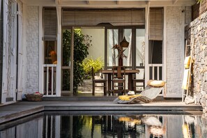A poolside area with a lounge chair, a table, and a chair, surrounded by a stone wall and a herringbone-patterned railing.