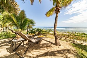 A beachside lounge with a view of the ocean, palm trees, and a sandy area with lounge chairs and a table.