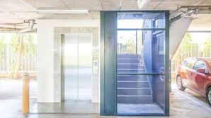Ein Aufzug mit Glastür, eine Treppe, die zu einer oberen Ebene führt, und ein rotes Auto, das draußen geparkt ist.