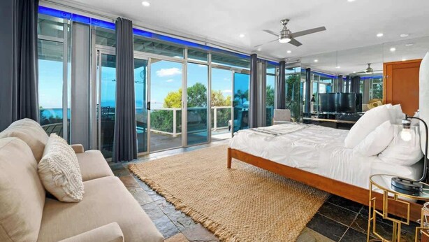 A spacious bedroom with a large bed, a sofa, and a TV. It features a sliding glass door leading to a balcony with a view of the ocean.