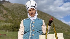 Een persoon in traditionele kledij, een tas vasthoudend, staat in een grasveld met bergen op de achtergrond.
