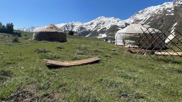 Een joert met rieten dak en een grote, ronde, witte tent in een grasveld, met bergen op de achtergrond.