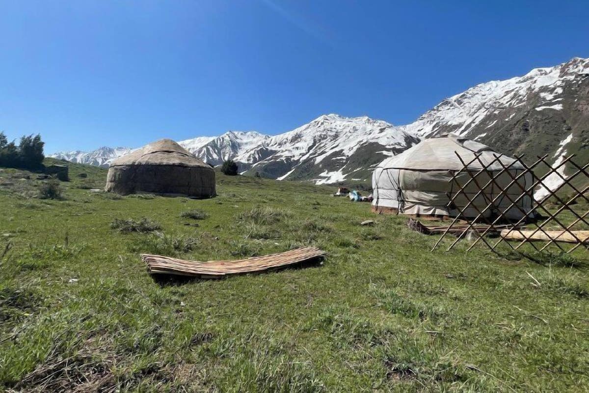 A yurt with a thatched roof and a large, round, white tent in a grassy field with mountains in the background.