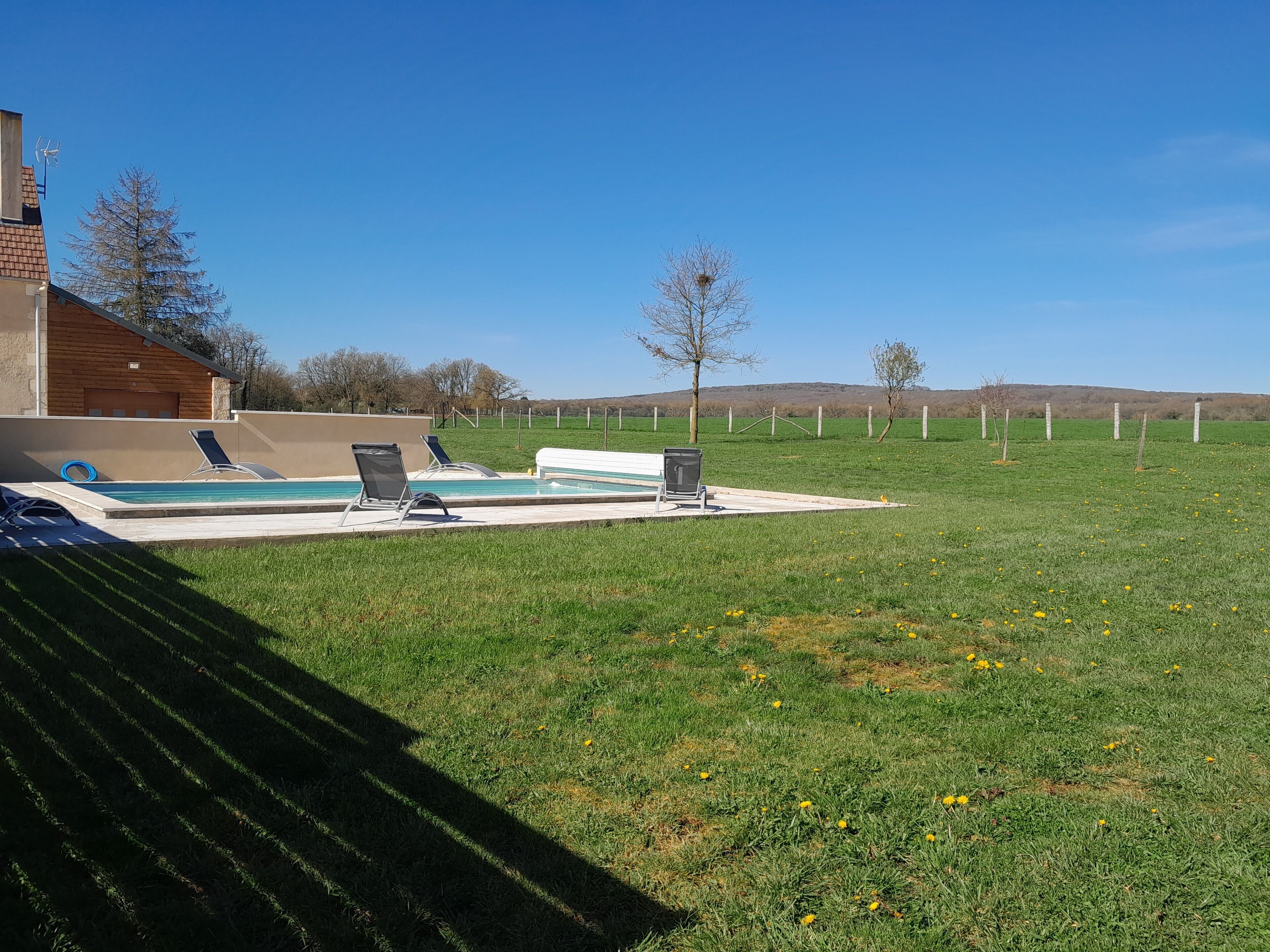 Una piscina con sillas de descanso, un banco y una zona verde con árboles y colinas al fondo.