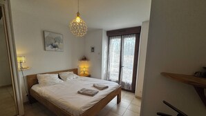 A bedroom with a wooden bed, a hanging light fixture, a bedside table with a lamp, and a window with curtains.