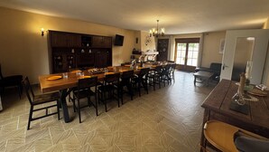 A spacious dining area with a long wooden table, multiple chairs, and a kitchen area with cabinets and a chandelier.