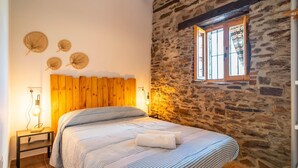 A bedroom with a wooden headboard, a stone wall, a window with curtains, and a bedside lamp.