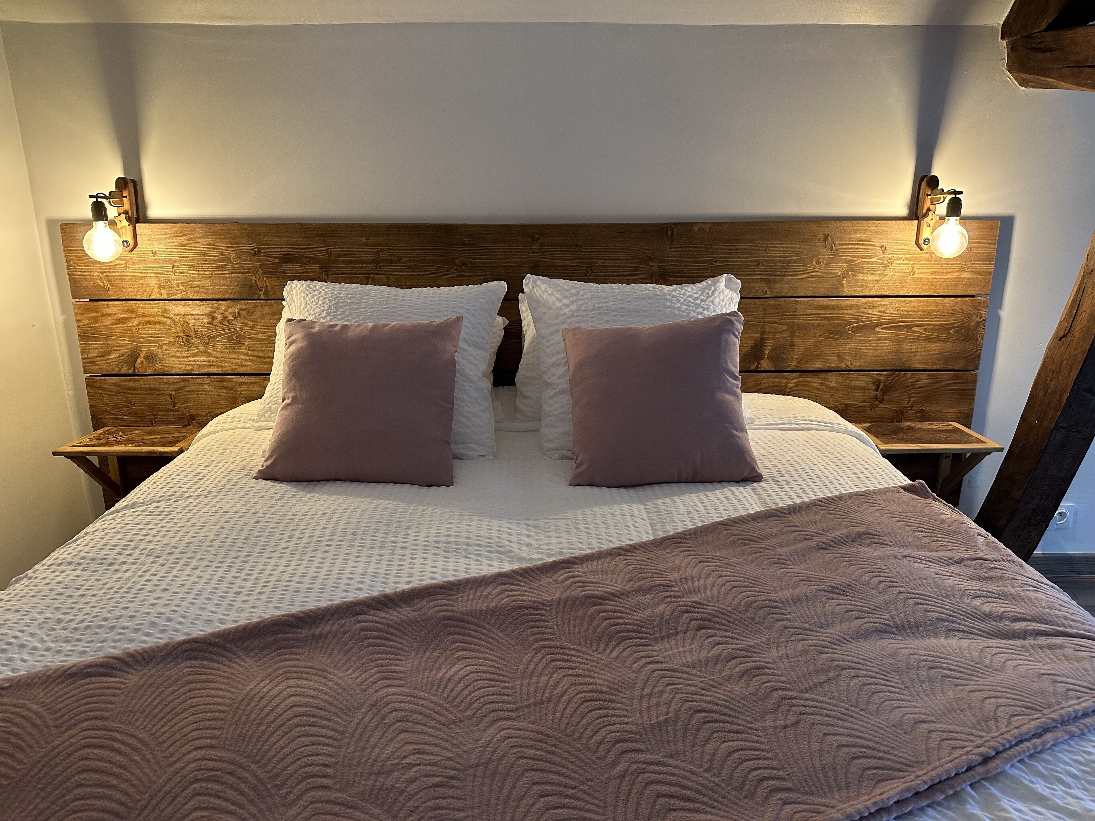 Uma cama bem arrumada com cabeceira de madeira, duas mesas de cabeceira e duas luminárias fixadas na parede.