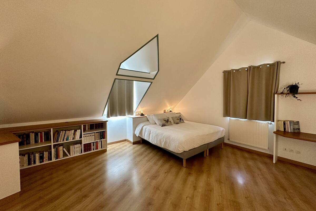 A bedroom with a sloped ceiling, a bed, a bedside table with a lamp, a bookshelf, and a window with curtains.