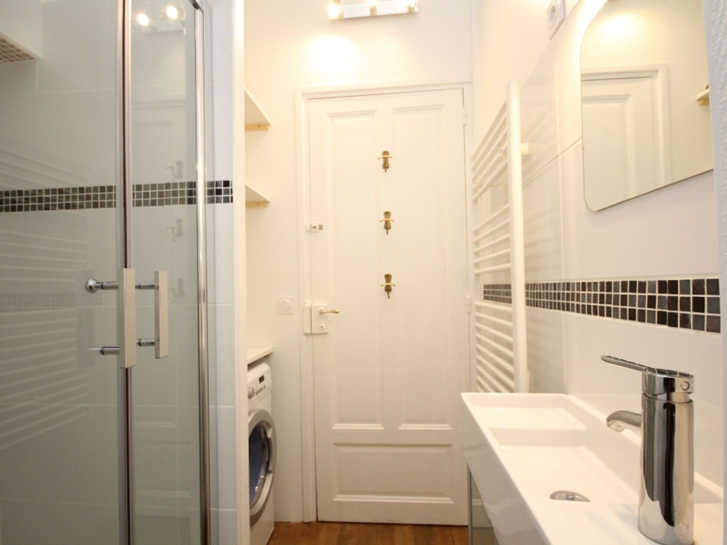 A bathroom with a glass shower enclosure, a white door, a washing machine, and a sink with a chrome faucet.